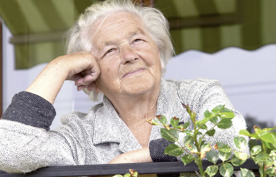 Ältere Frau sitzt auf dem Balkon, hat den Kopf auf die Hand gestützt und schaut zufrieden in die Ferne